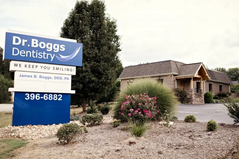 A large outdoor sign displays "Dr. Boggs Dentistry" along with the tagline "We Keep You Smiling!" and contact details. Behind the sign, a dental office building with a stone facade and wooden entrance is surrounded by well-maintained landscaping.