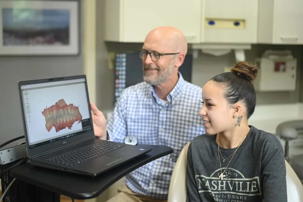 Two individuals in a dental office, with one seated in a dental chair and Dr. Boggs pointing at a laptop displaying a 3D dental model, illustrating digital technology use in dental practices.