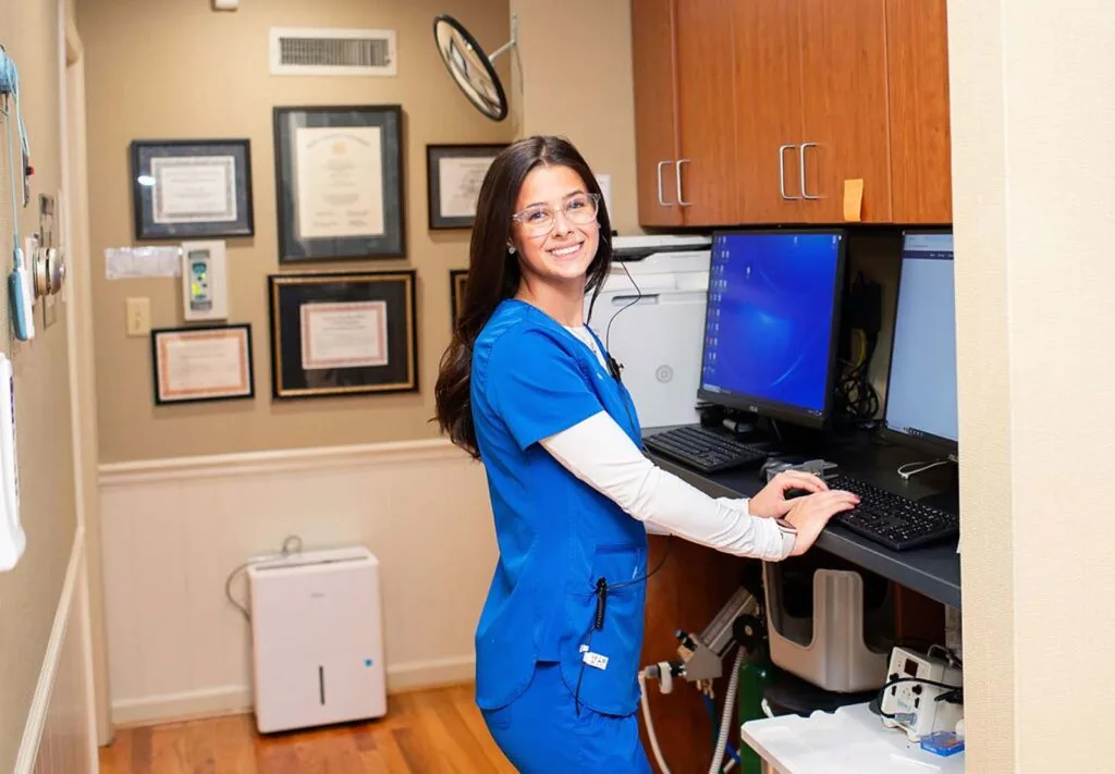 A person wearing blue scrubs stands at a desk in a medical office. The desk holds a computer with a blue screen, a printer, and other office equipment. Framed certificates adorn the wall behind them, and a white air purifier is on the floor.