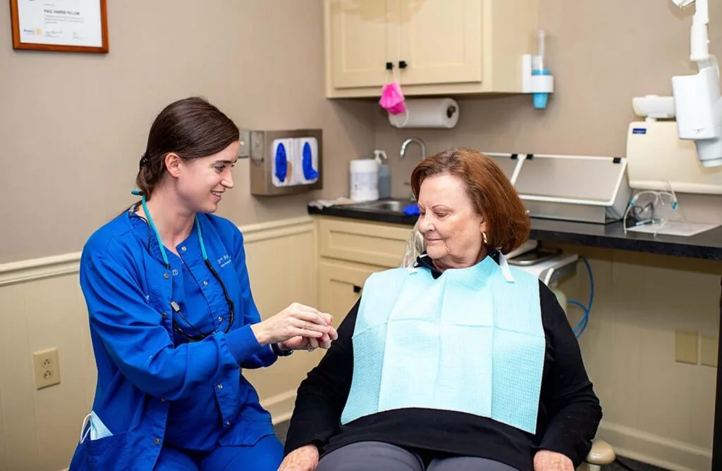 Dental professional in blue uniform interacts with a patient in a dental chair; background includes dental equipment, supplies, and a framed certificate on the wall.