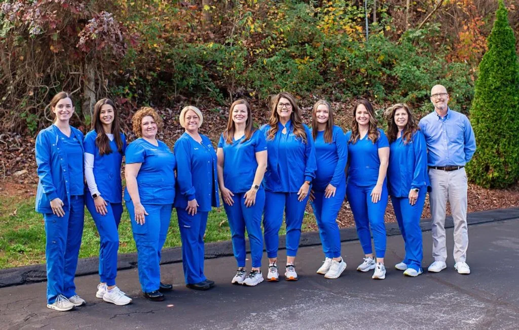 A group photo of 10 individuals standing on a paved surface outdoors with greenery in the background. Nine wear blue medical scrubs, and one on the far right wears a light blue button-up shirt and light-colored pants, suggesting they might be healthcare professionals.