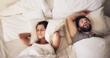 A man snores loudly while sleeping on his back as a woman beside him covers her ears with a pillow, illustrating disrupted sleep.